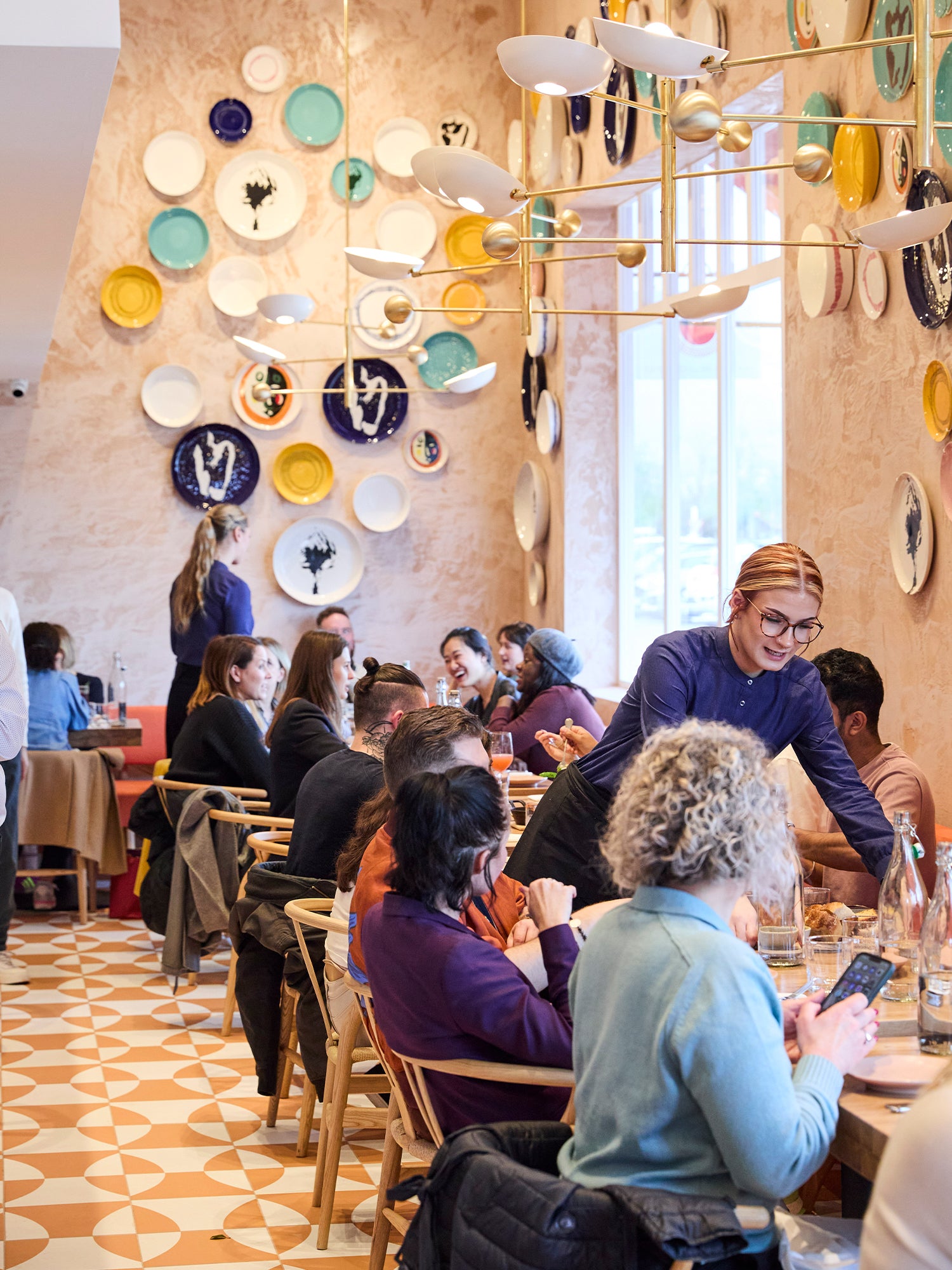 Dining room with a display of ceramics on the wall. Diners at the table, waiters serving.
