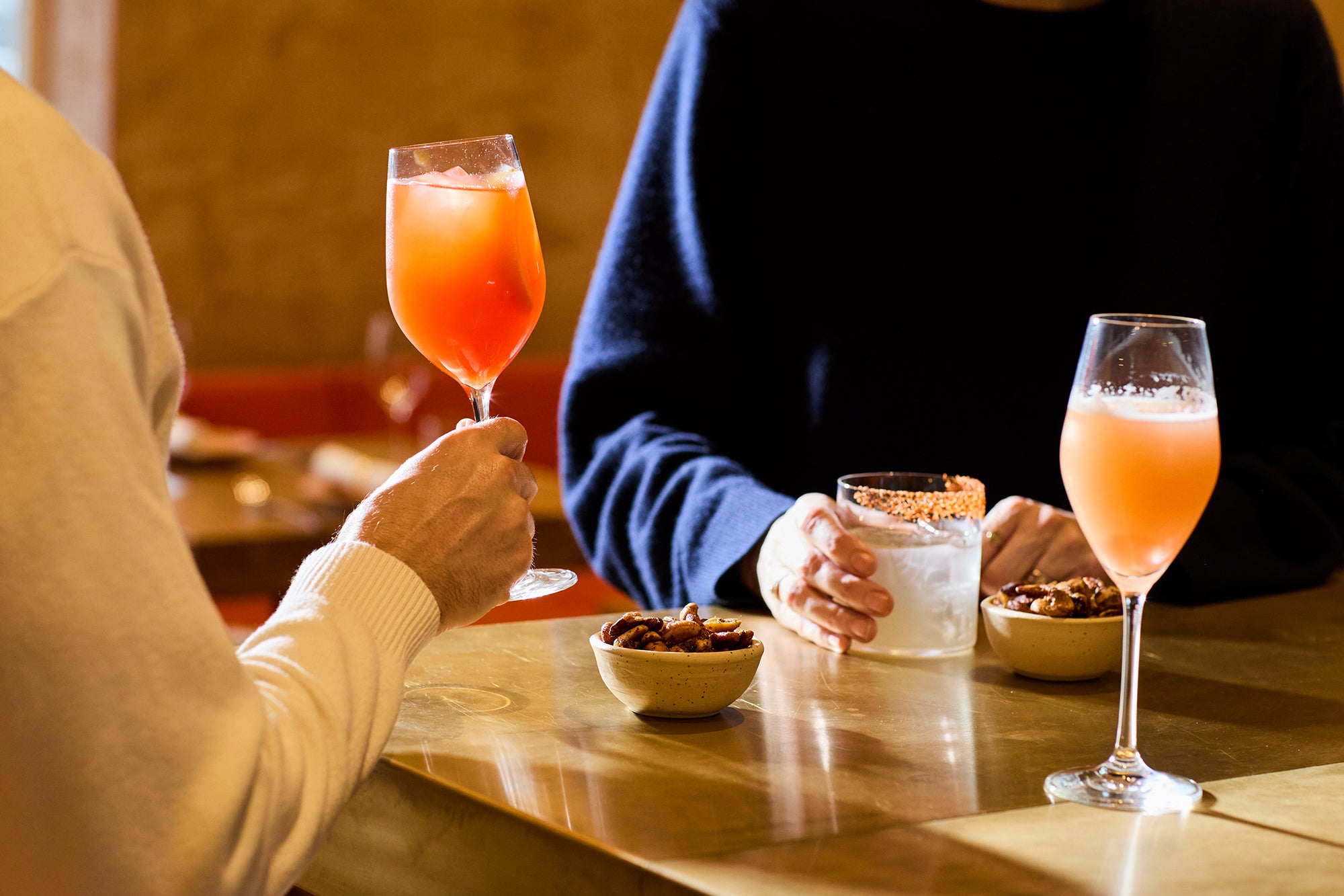 Three glasses with cocktails, people sharing rosemary spicy nuts.